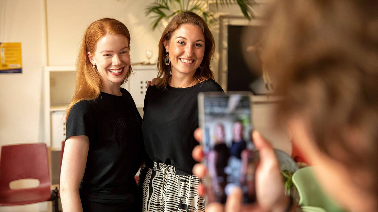Twee vrouwen die lachend kijken terwijl er met een telefoon een foto van hen wordt gemaakt.