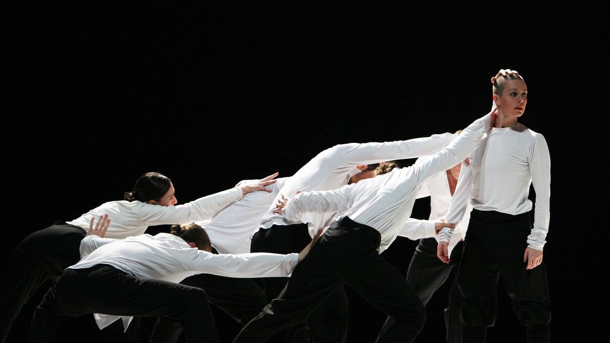 Zeven dansers in zwarte broeken en witte shirts, hangend tegen een staande persoon