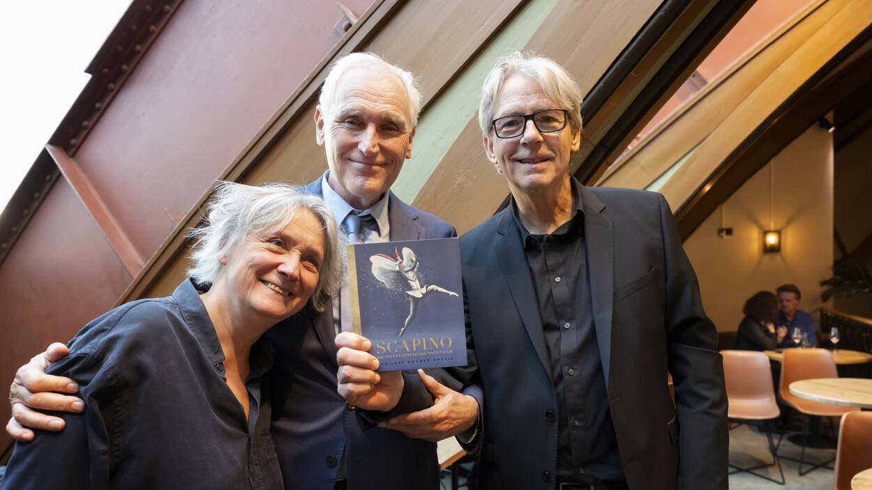Drie personen in nette kleding met tussen hen in een boekje met mooie illustratie van een springende danser en de titel ‘Scapino'.
