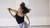 Danseres met donker halflang haar dat naar achter wappert in een lichte studio. Zij heeft een elleboog gebogen en schuin omhoog. Haar andere arm is naar achteren.